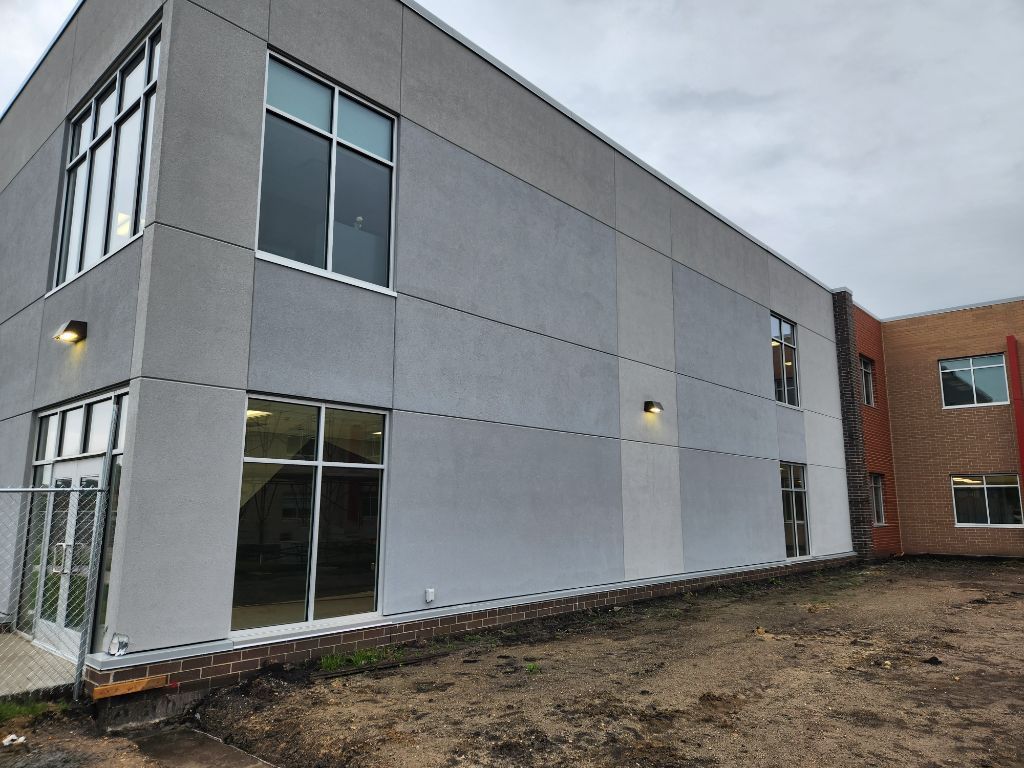 Modern gray building exterior with large windows beside a red brick wing and muddy ground