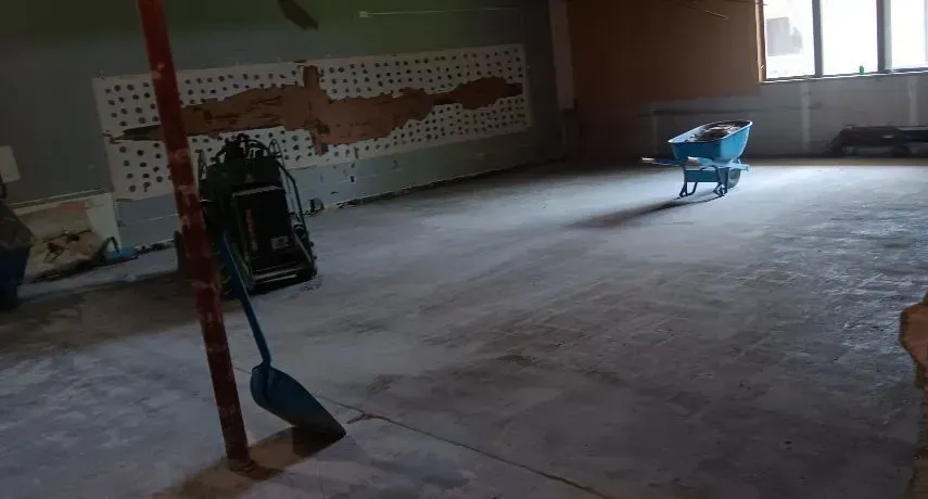 A construction site with a concrete floor, a blue wheelbarrow, and a floor sander in a room with a damaged wall.