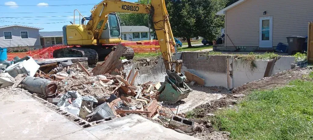 A yellow excavator demolishing a building foundation on a residential lot.