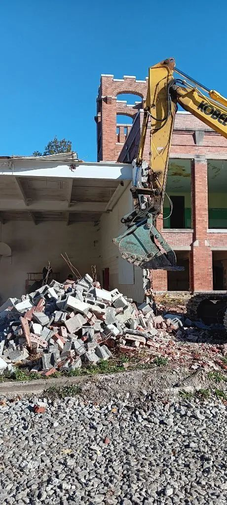 Yellow excavator demolishing a brick building, with rubble piled in front