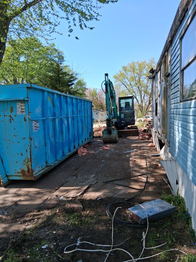 Blue dumpster and excavator beside a house on a dirt driveway under clear sky