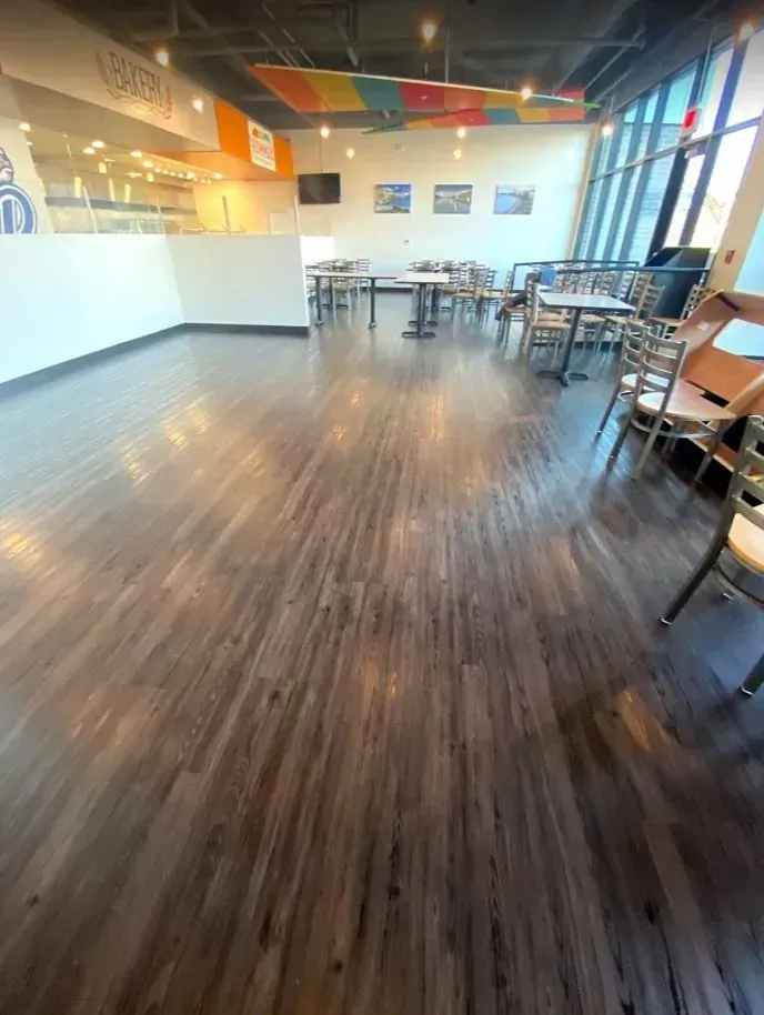 A brightly lit, modern dining area with wood-grain flooring, several tables and chairs, and a service counter in the back.