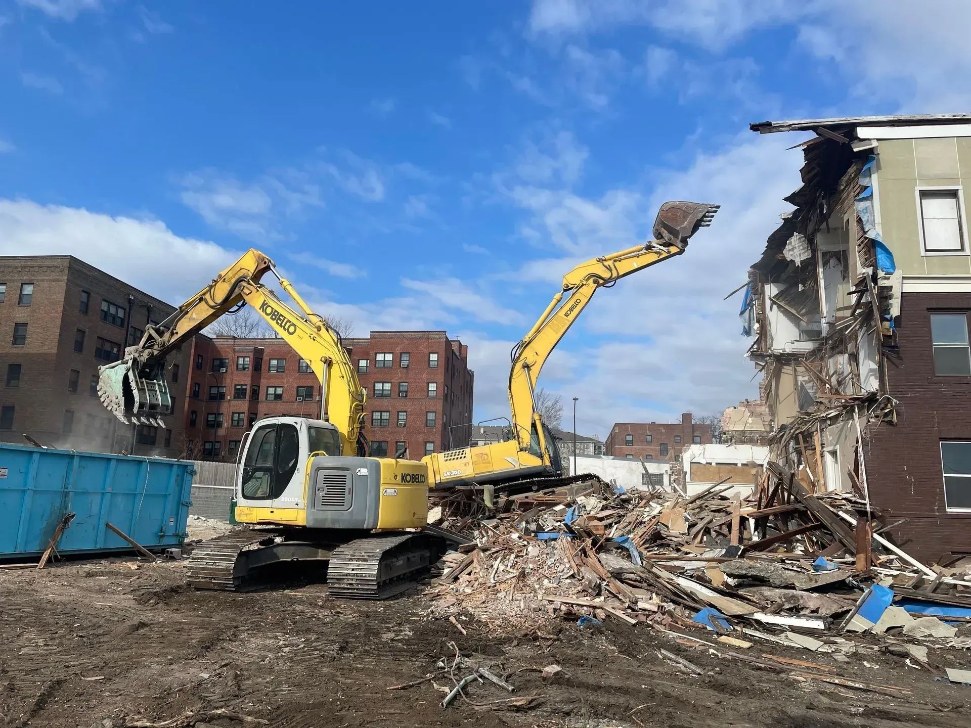 Yellow excavators demolishing a damaged brick building amid rubble under a blue sky.