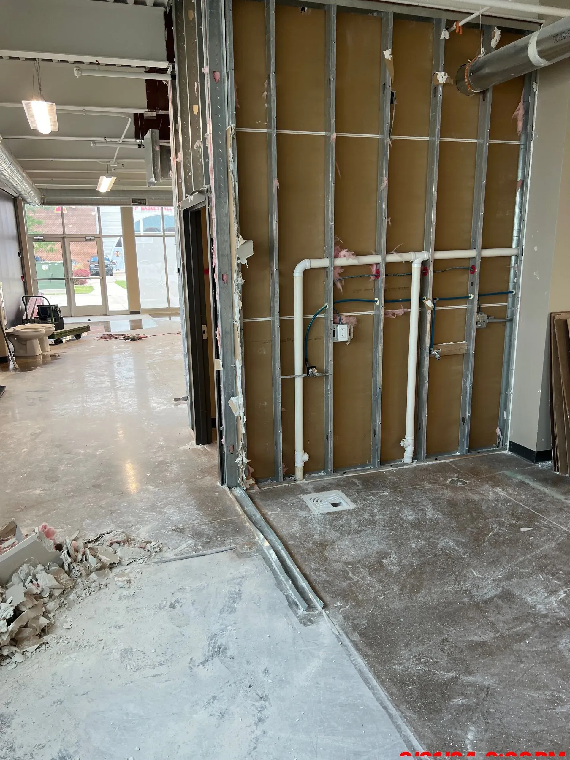 An interior view of a room under construction with exposed metal wall studs, plumbing pipes, and debris on the floor.