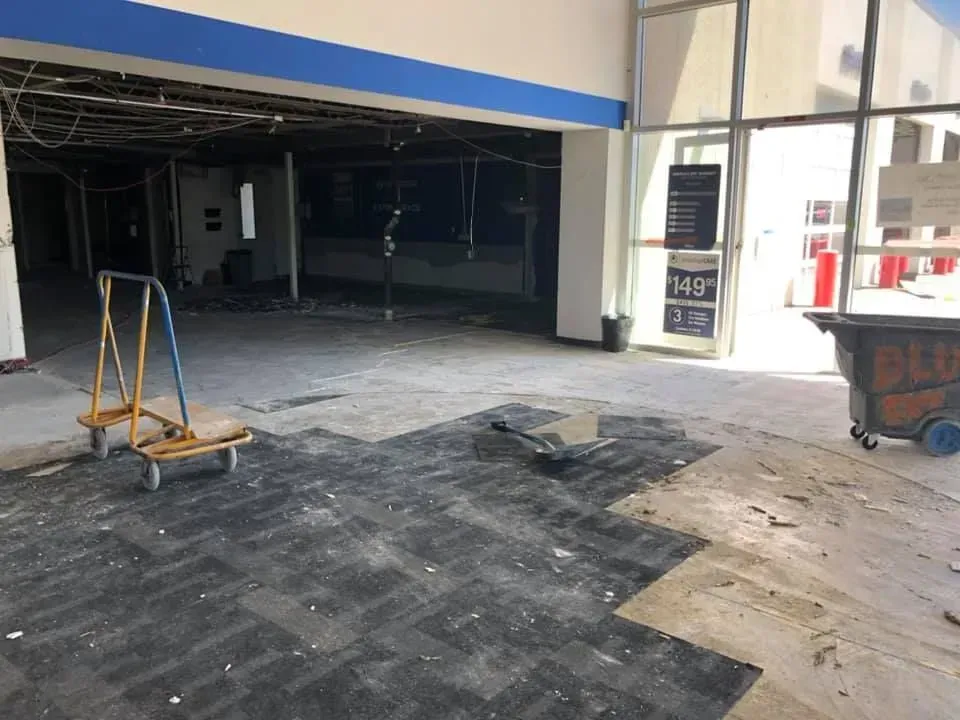 A construction site at a building entrance with a yellow flatbed cart, bare floor patches, and a large metal dumpster.
