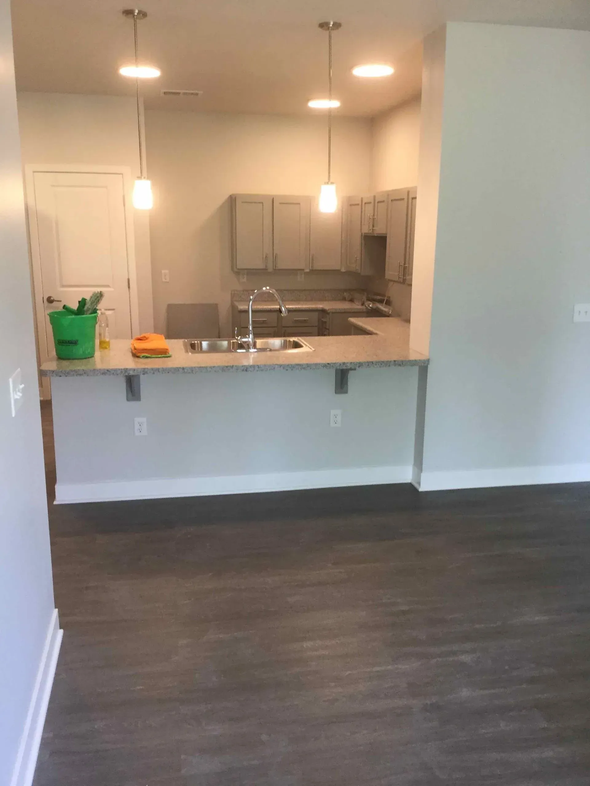 A modern kitchen area with light gray cabinets, a countertop peninsula, two pendant lights, and dark wood-look flooring.
