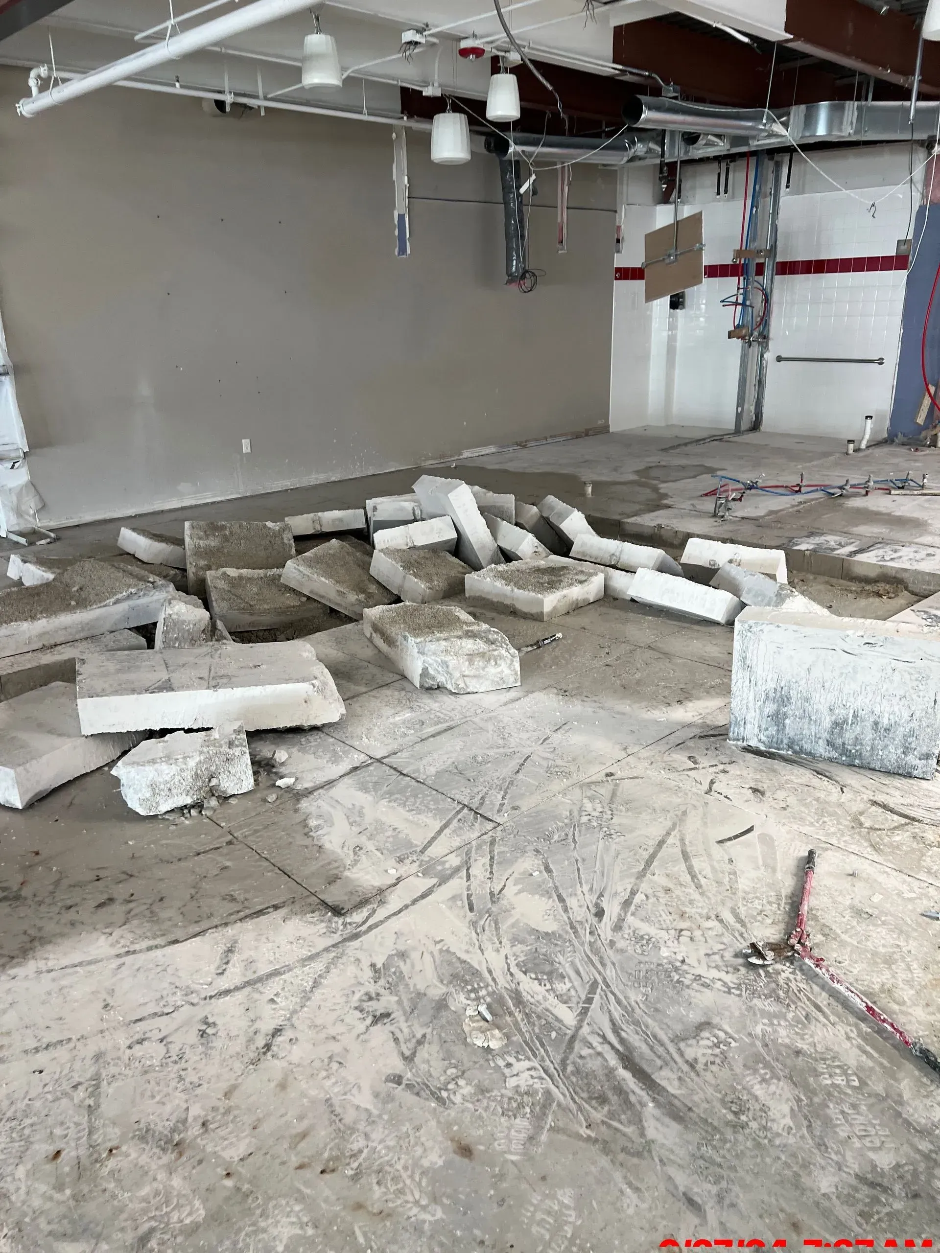 A construction site interior with scattered concrete debris and exposed overhead pipes against a plain, light-colored wall.