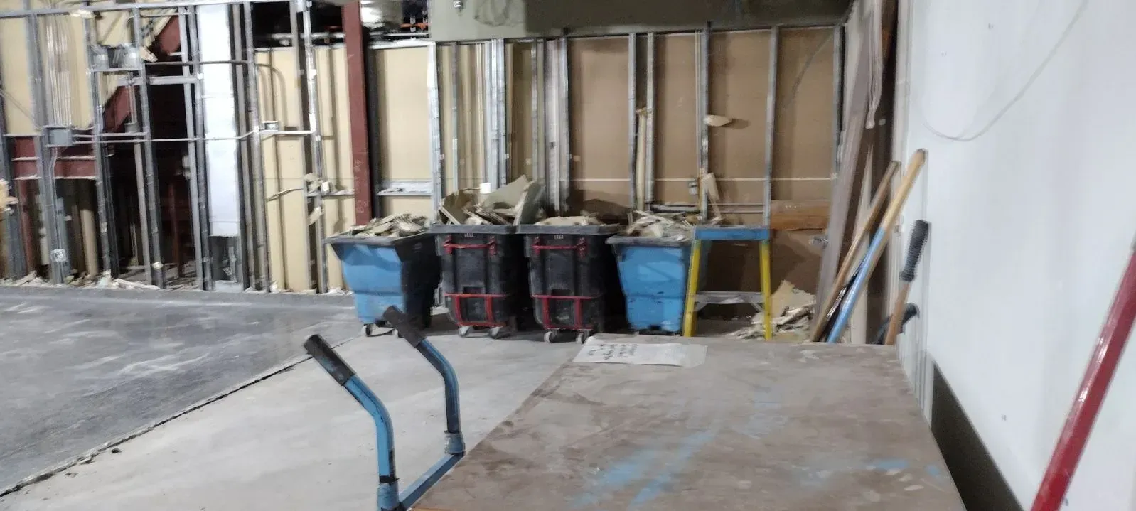 Construction zone with metal wall framing, four blue debris bins, a workbench, and scattered tools in an indoor space.