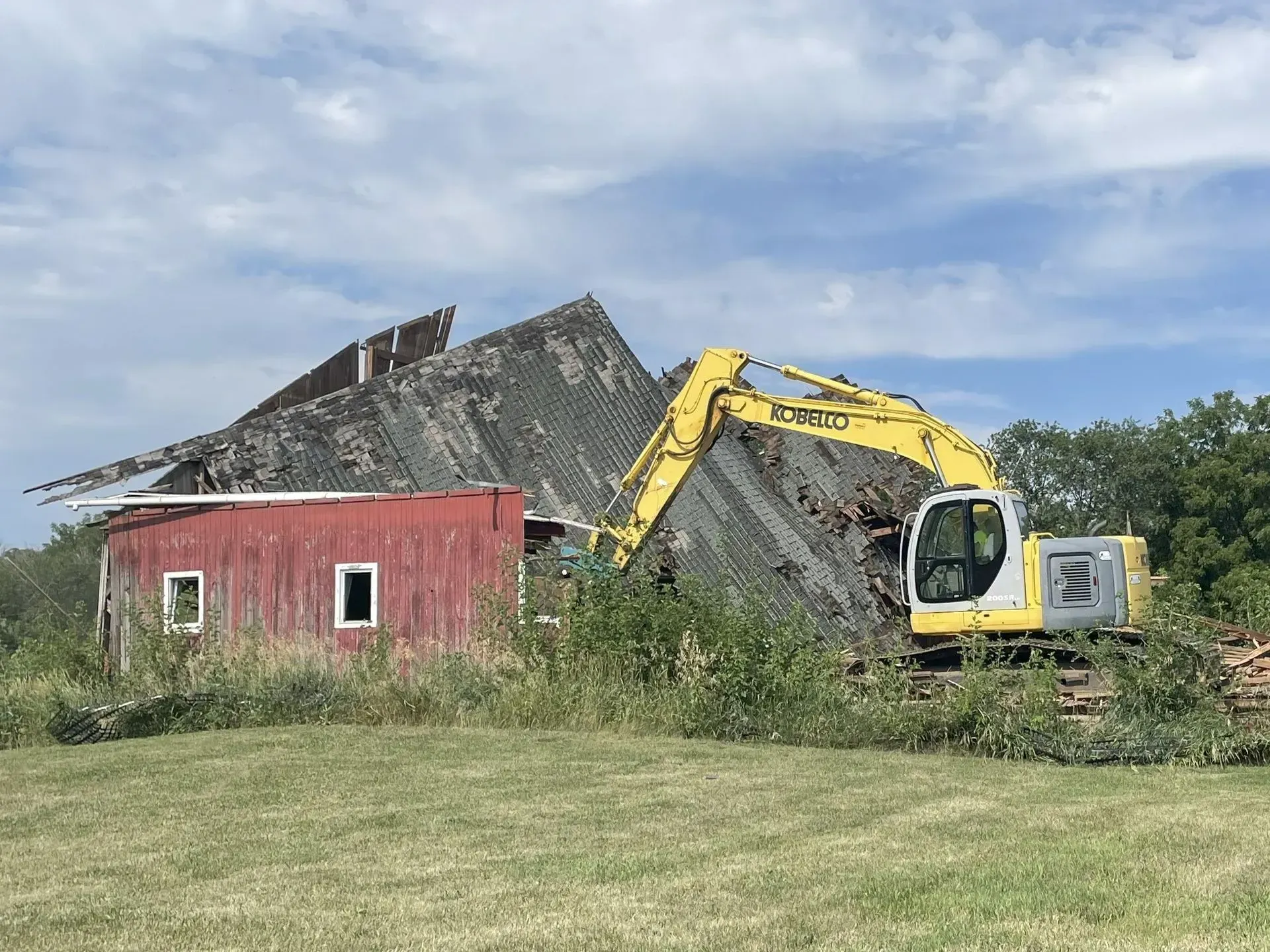 A yellow excavator demolishes a dilapidated barn with a red side wall in an open field under a cloudy blue sky.