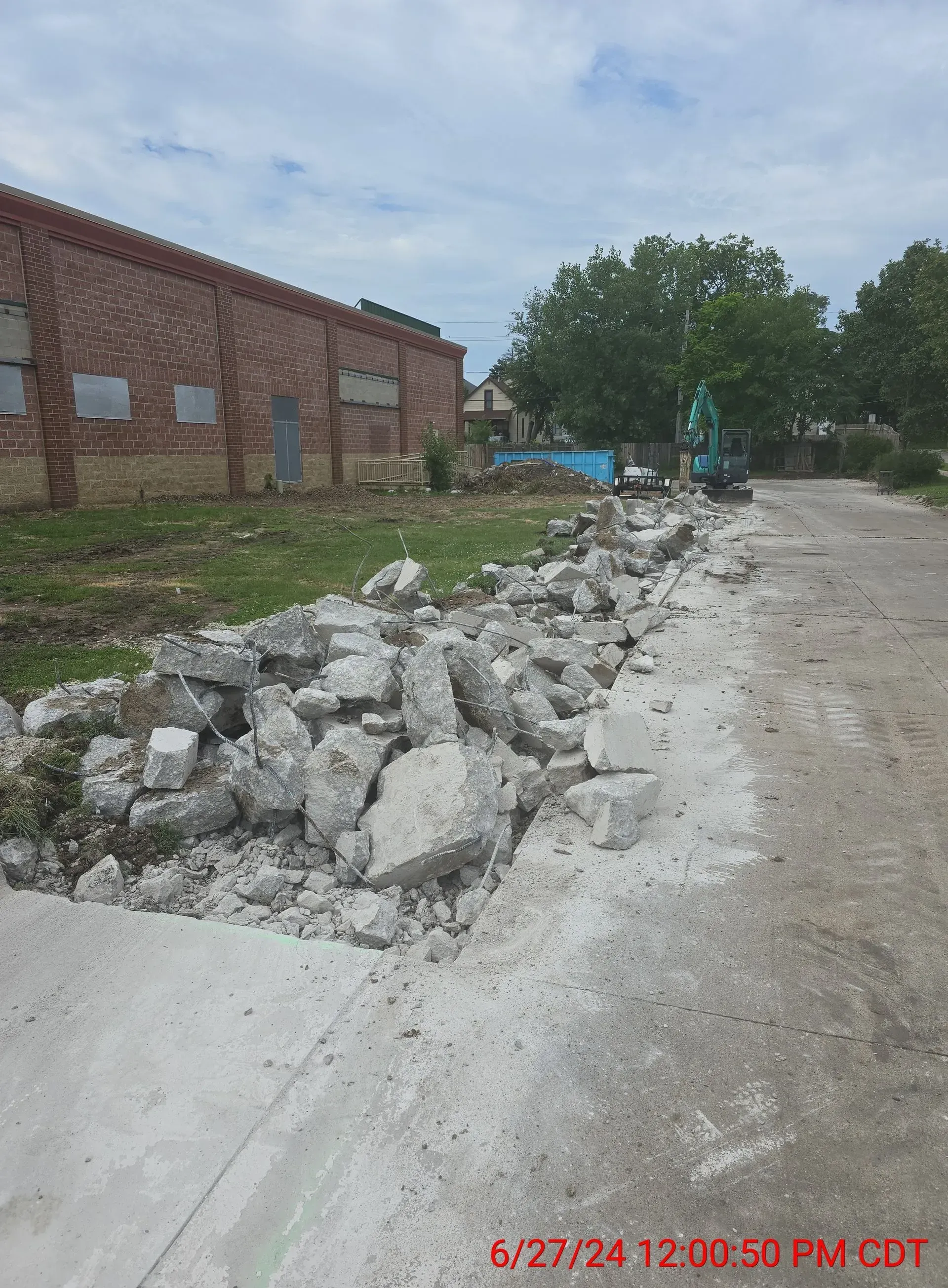 Construction debris and broken concrete piled along the edge of a brick building near a gravel path under a cloudy sky.