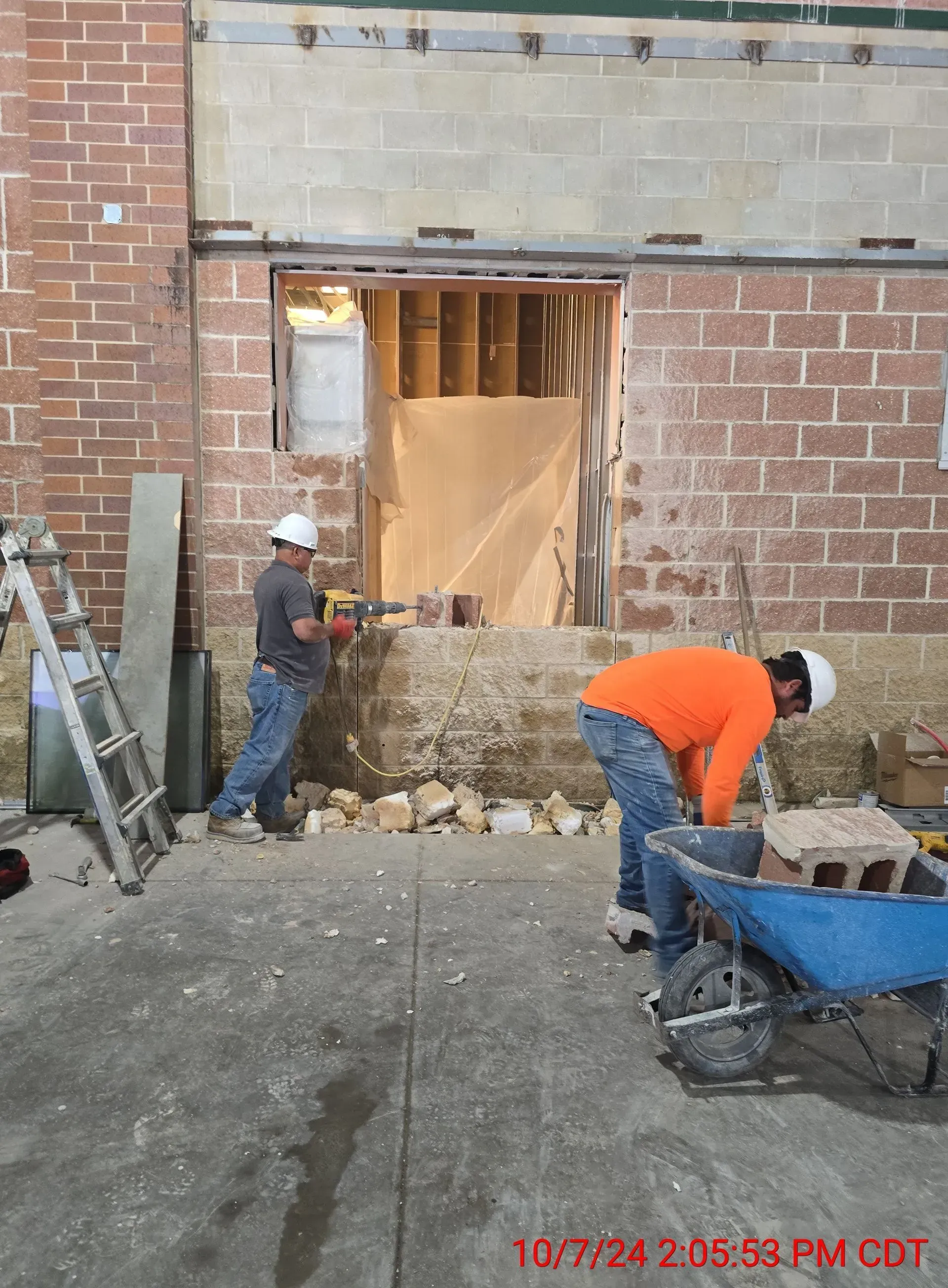 Two workers in hard hats and high-visibility gear renovate a brick wall opening, with one using a tool and one with a cart.