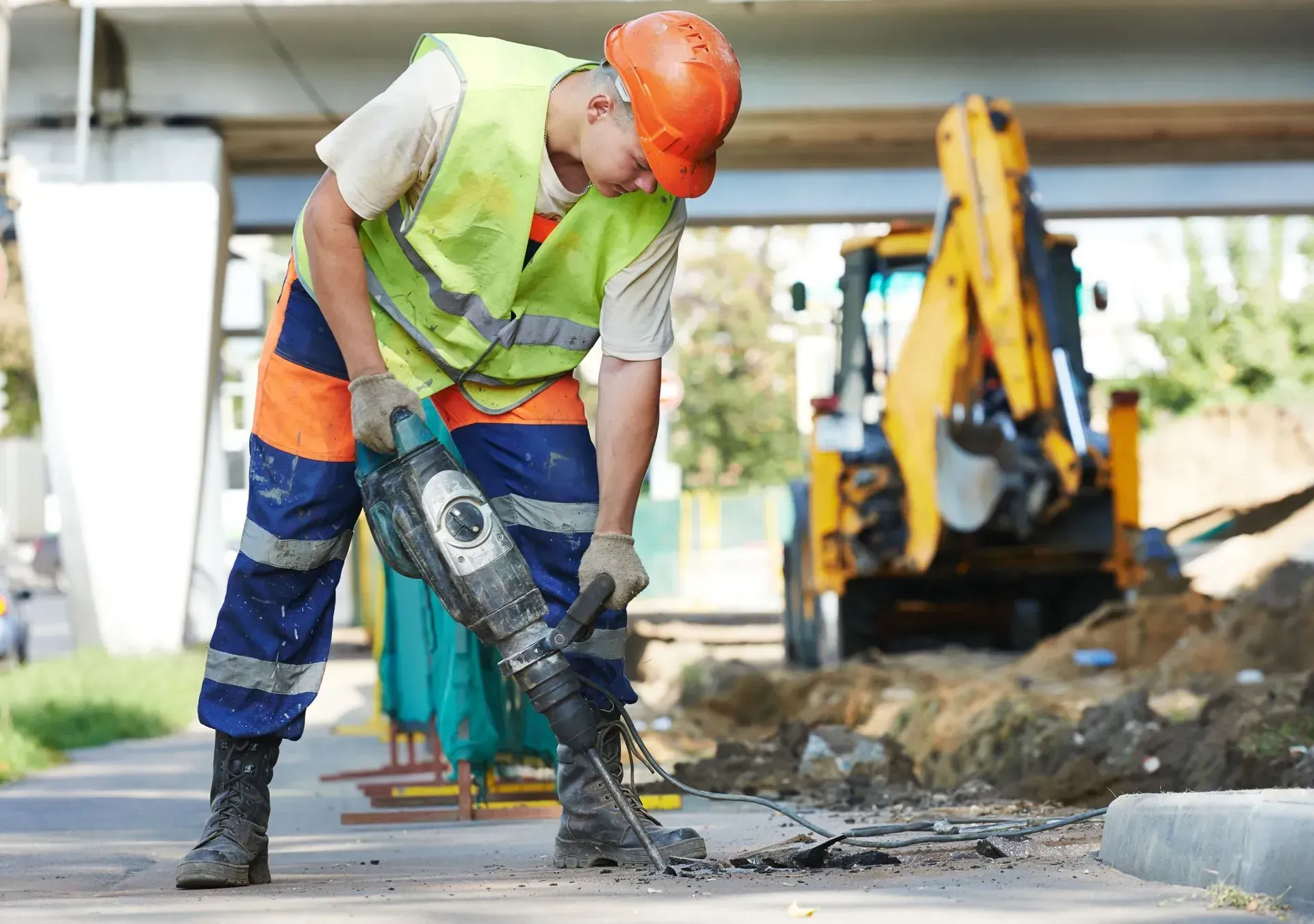 A construction worker in a high-visibility vest and orange hard hat uses a jackhammer on a sidewalk near a yellow excavator.