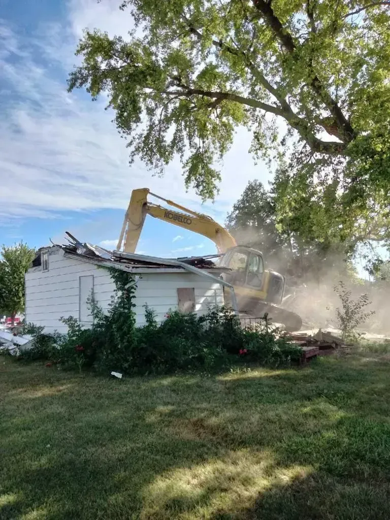 A yellow excavator tears down a small, white house surrounded by grass and trees on a sunny day.