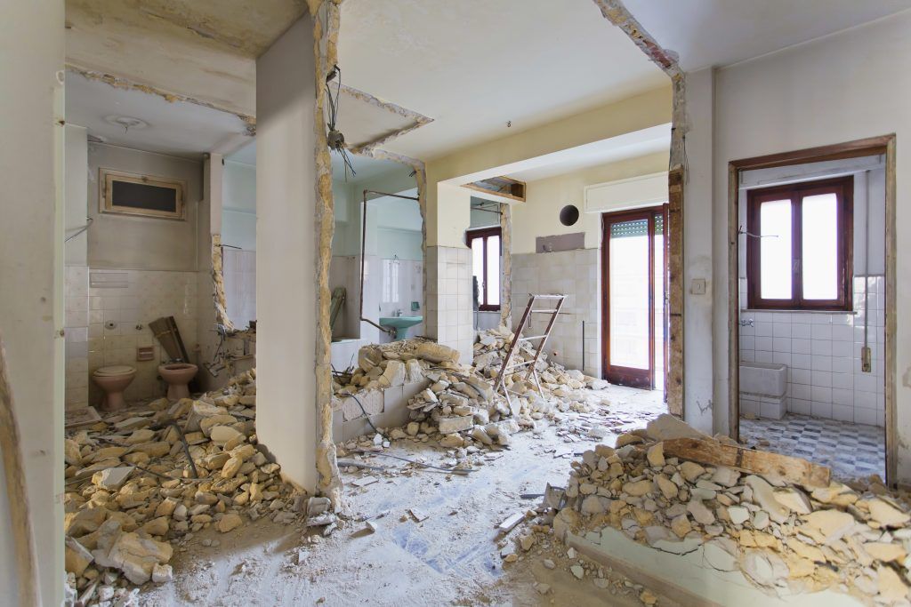 An orange demolition machine tears down a section of a multi-story brick building, leaving exposed rubble.