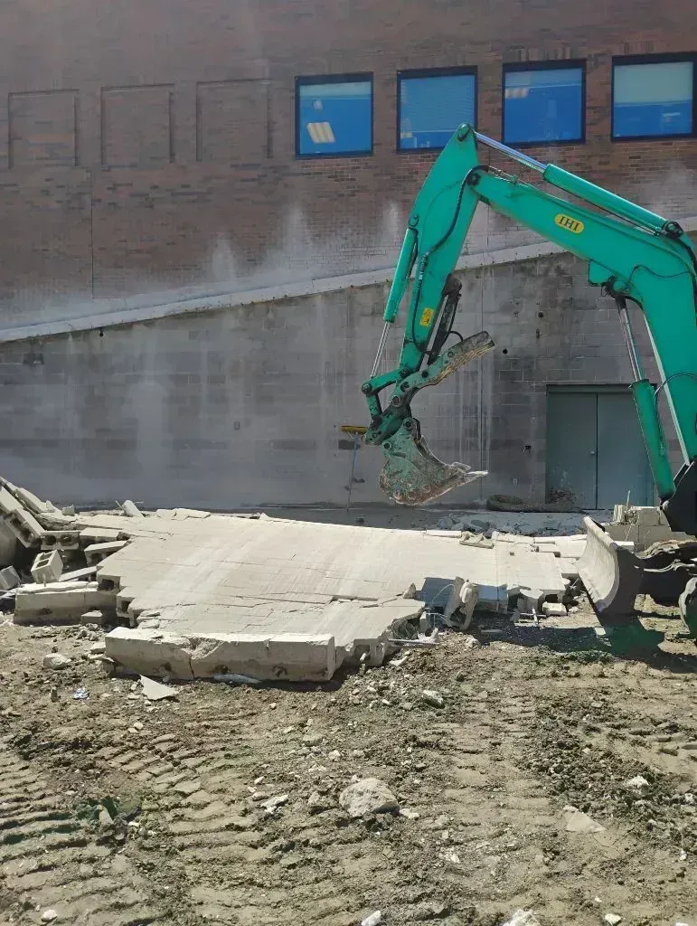 Green excavator breaking concrete beside a brick wall on a construction site