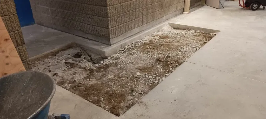 Construction scene showing a rectangular section of concrete flooring removed, revealing the gravel base near a wall.