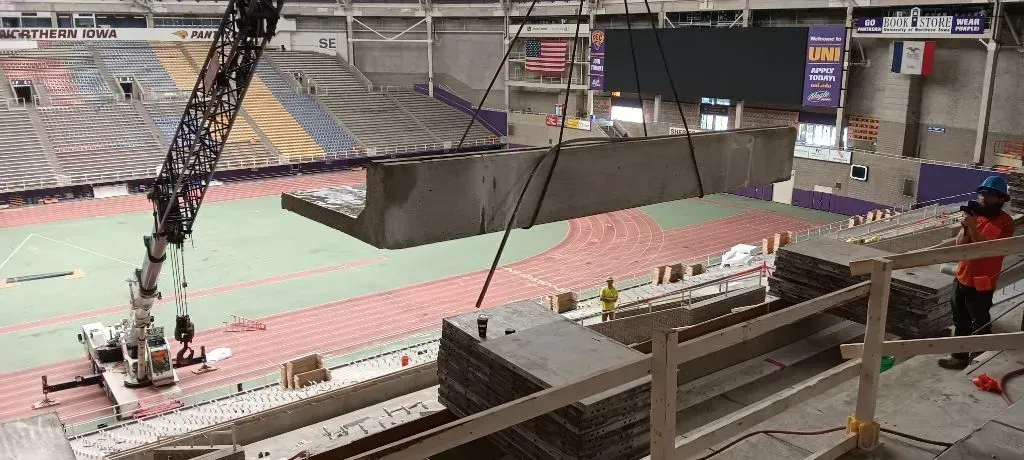 Indoor arena with a large crane lifting a dark panel near a track and stadium seating