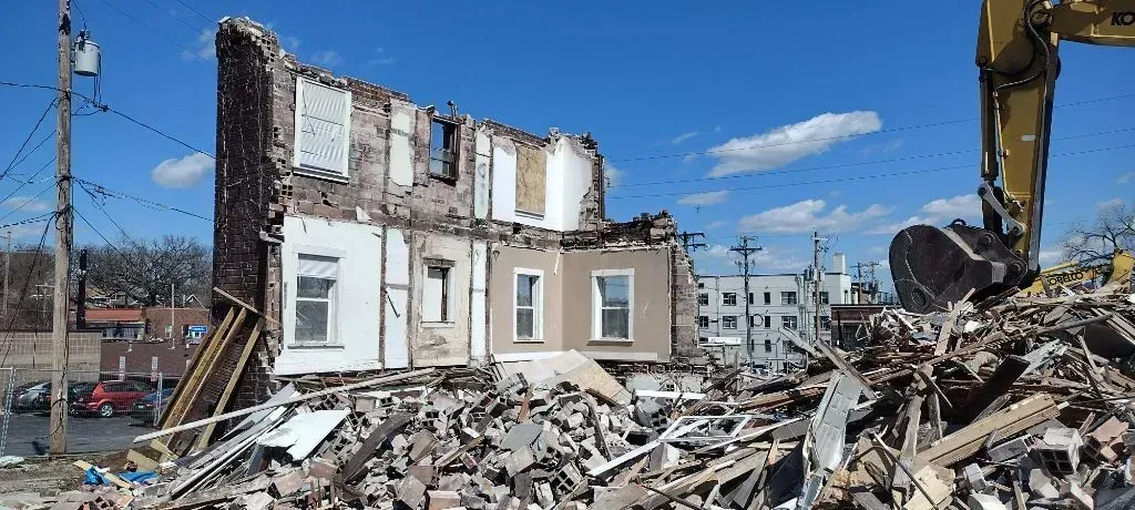 A large yellow excavator demolishes a multi-story building, leaving behind a partially standing facade and debris.