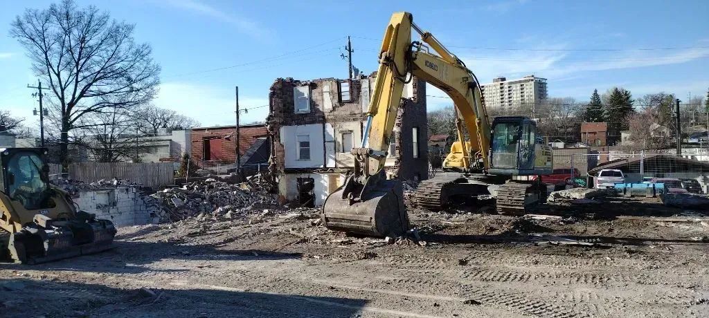 Excavator demolishing a building at a construction site amid rubble and parked cars.