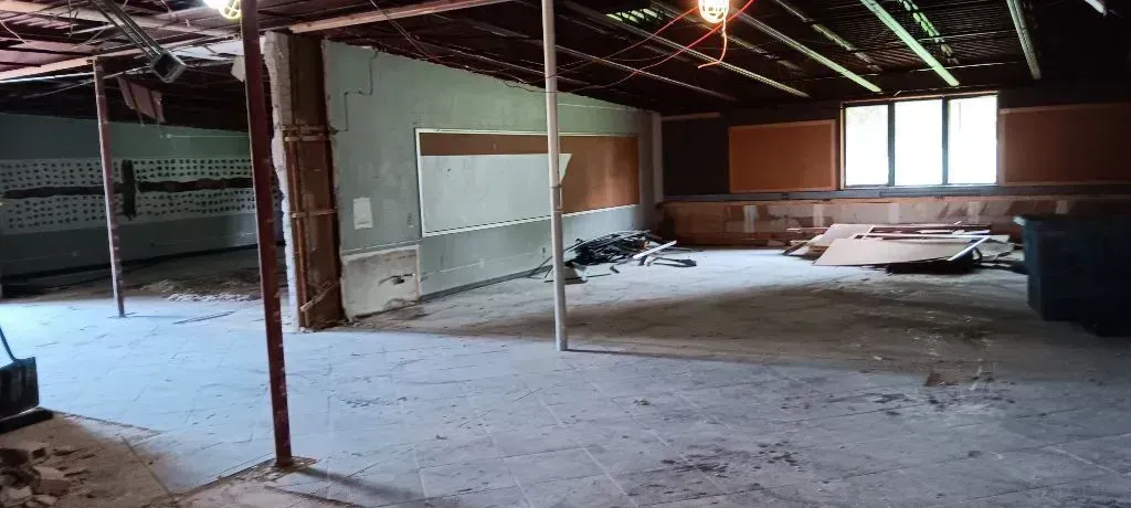 Abandoned empty room with exposed beams, debris on floor, and windows letting in daylight
