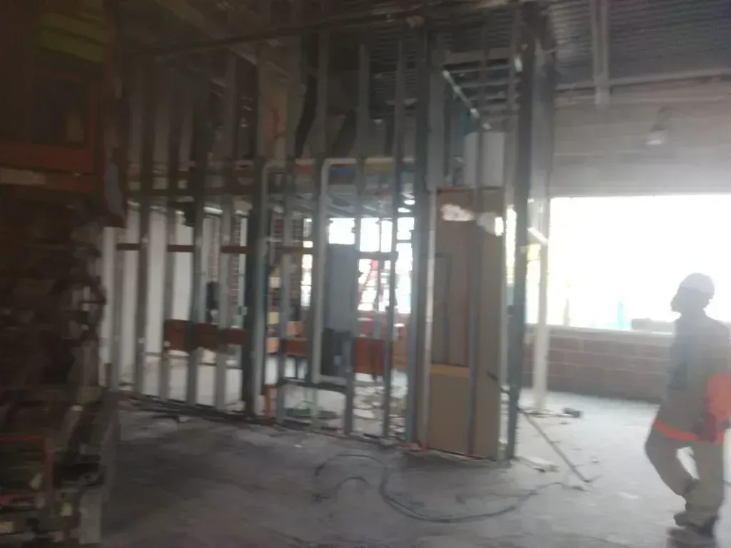A construction worker in a high-visibility vest stands in an unfinished room with metal wall framing and ductwork.