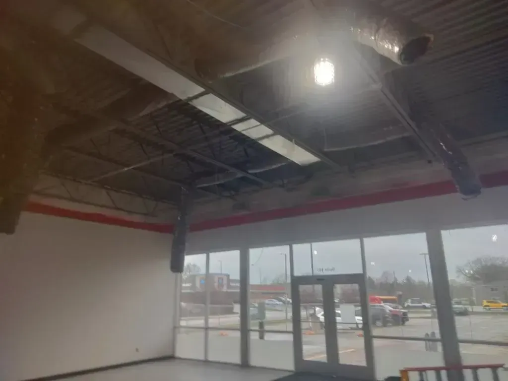 Interior of an empty commercial space with high ceilings, exposed ductwork, a single light, and floor-to-ceiling windows.