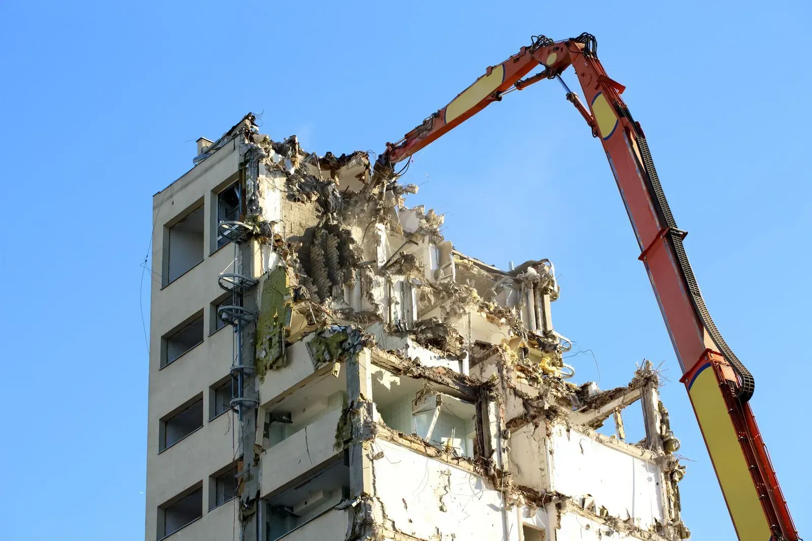 A high-reach excavator arm tears down the top of a multi-story concrete apartment building against a clear blue sky.