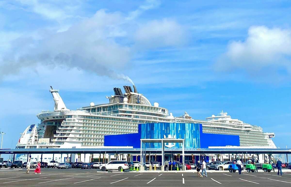 Large white cruise ship docked at a port under a blue sky, with people and cars in front.