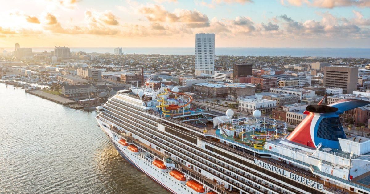 Large cruise ship docked by a city waterfront at sunset, with calm water and a tall tower in the distance