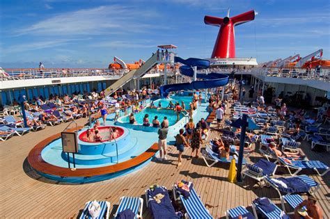 Crowded cruise ship pool deck with blue loungers, a hot tub, and a red funnel under a clear sky