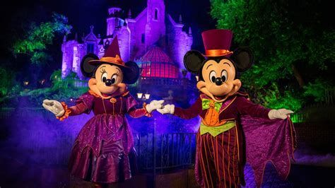 Mickey and Minnie in purple Halloween costumes pose in front of a glowing purple castle at night