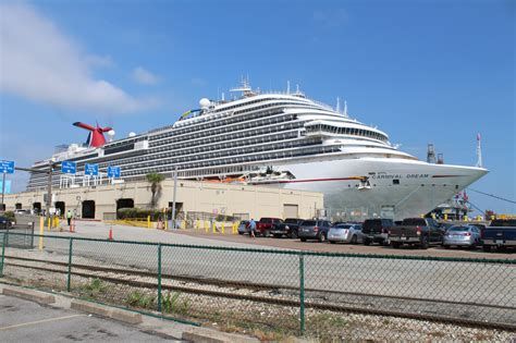 Large white cruise ship docked at a port beside a chain-link fence under a blue sky