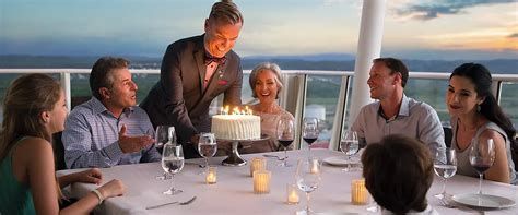 Friends dining on a cruise ship deck at sunset, with a waiter serving a candlelit table.
