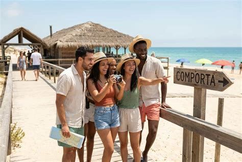 Four friends posing by a beach boardwalk sign reading “Comporta” on a sunny seaside pier.