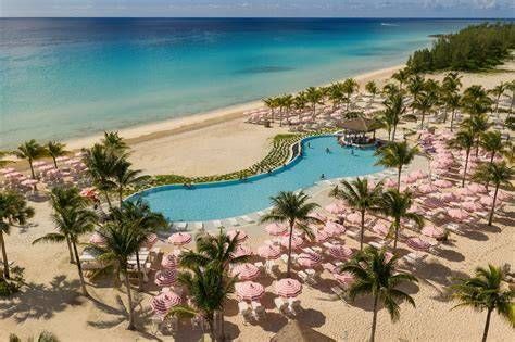 Tropical resort with a winding blue pool, pink umbrellas, palm trees, and a sandy beach by turquoise water