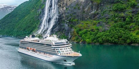 White cruise ship sailing beside a green cliff and waterfall in a calm fjord