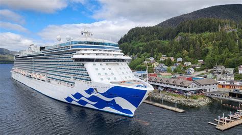 Large white cruise ship with blue wave design docked by a forested coastal town and hillside.