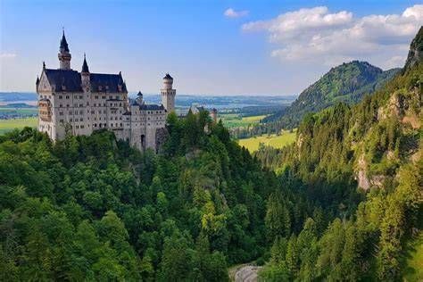 Neuschwanstein Castle above a lush green forest valley under a blue sky with clouds