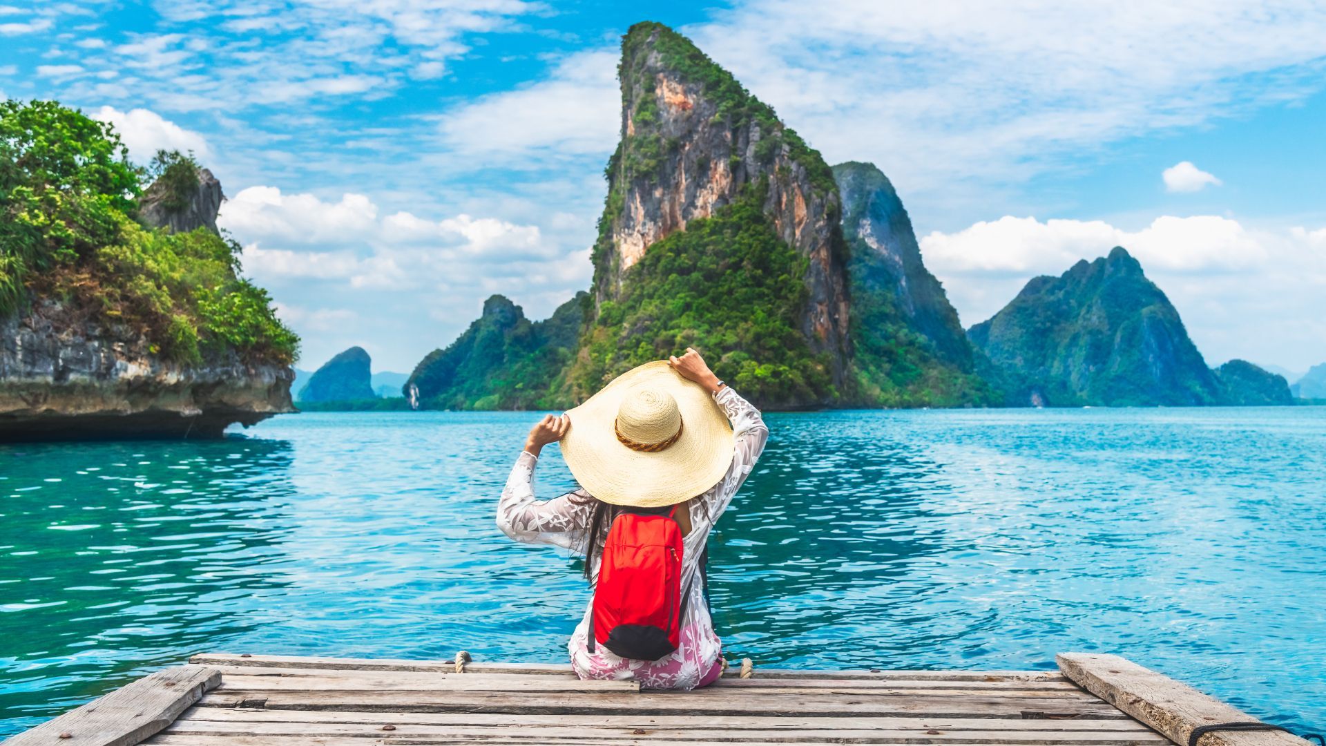 A person in a straw hat sits on a dock overlooking turquoise water and a rocky tropical island.