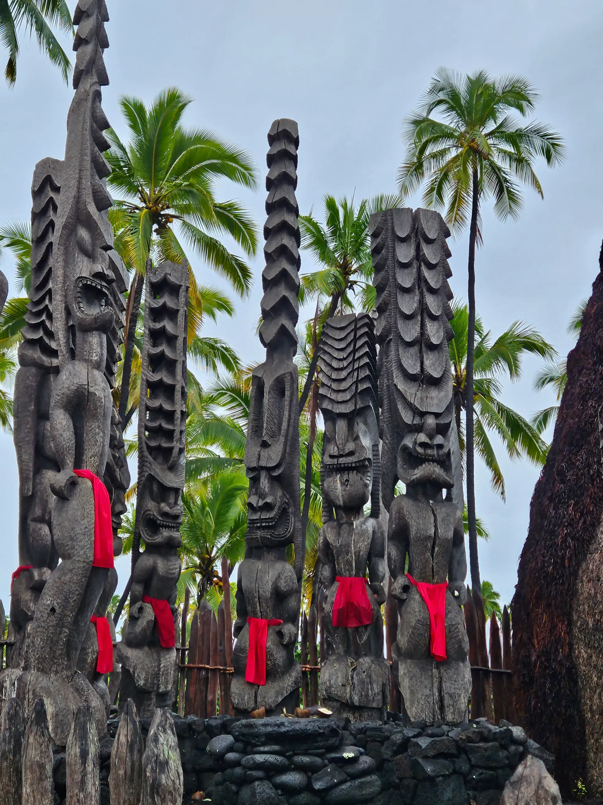 A group of totem poles with red ribbons around them are surrounded by palm trees.