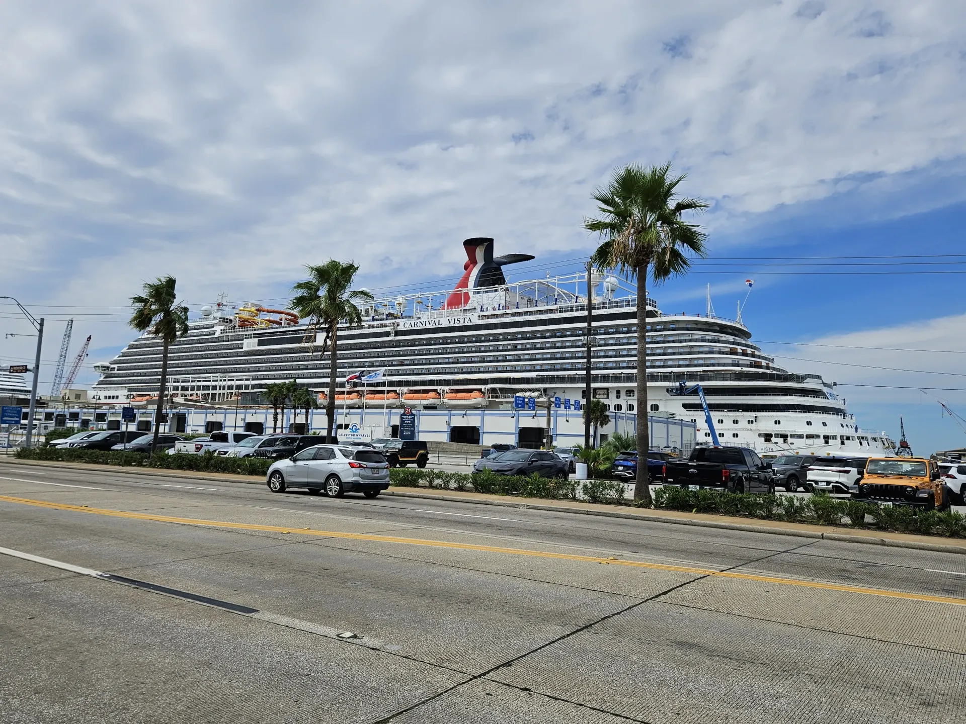 A large cruise ship is docked in a harbor with cars parked in front of it.