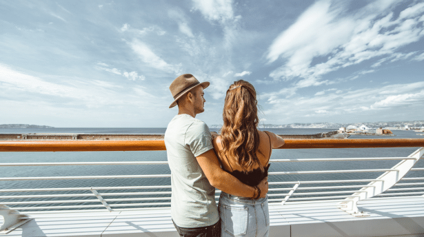 Two people stand on a cruise ship deck, looking out over the ocean under a bright sky.