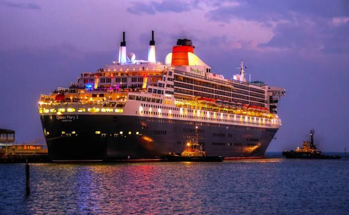 Large cruise ship lit up at dusk on calm water, with a purple sky and small tugboats nearby.