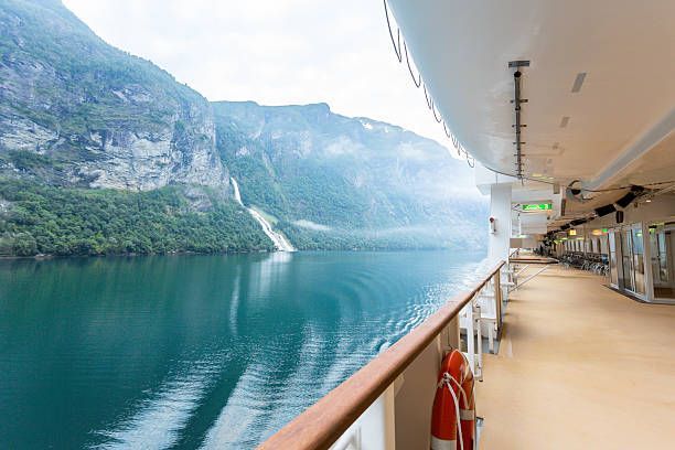 Cruise ship deck beside calm blue fjord and steep green cliffs under a cloudy sky
