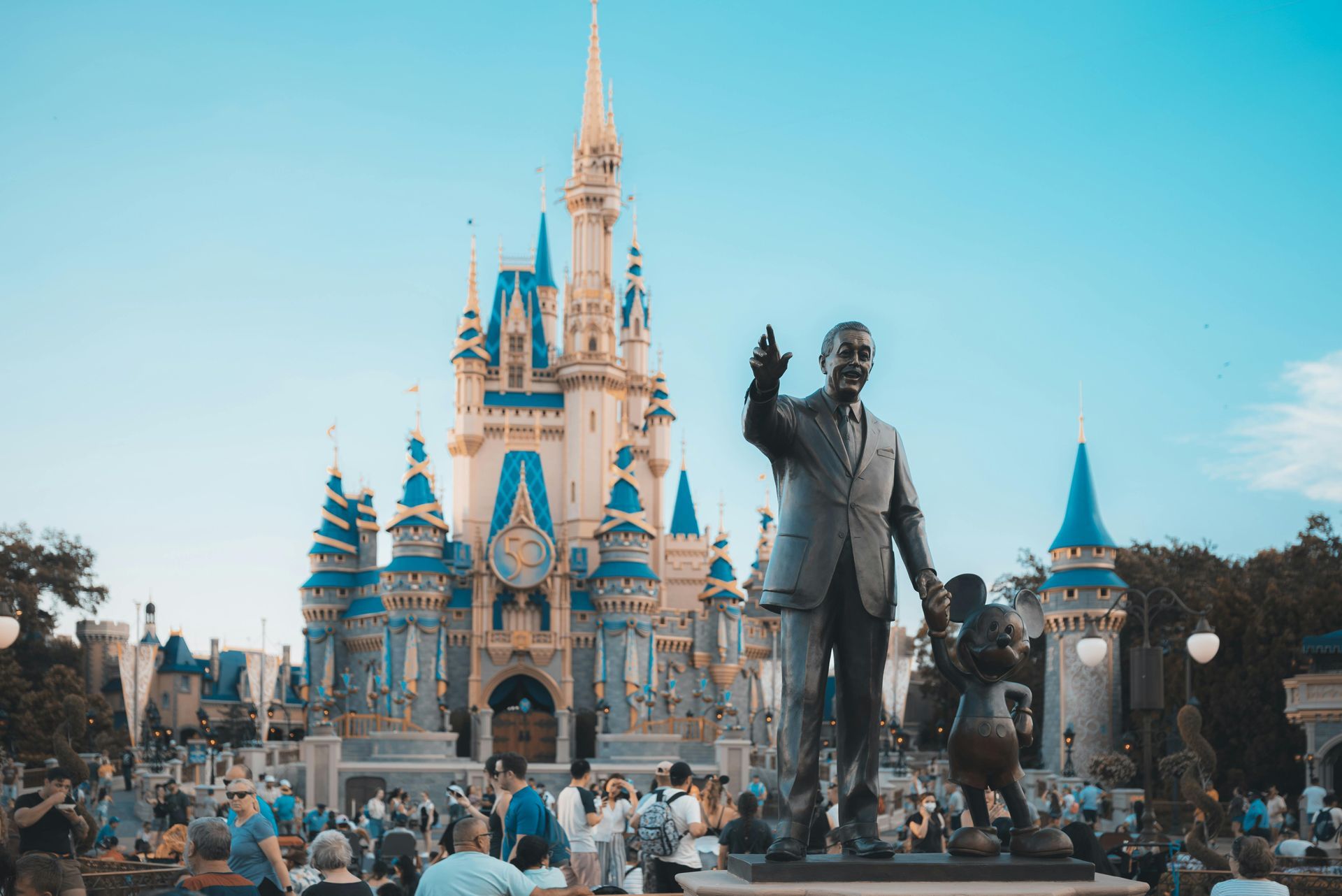 Cinderella Castle with a bronze Walt Disney statue and crowd in front under a blue sky