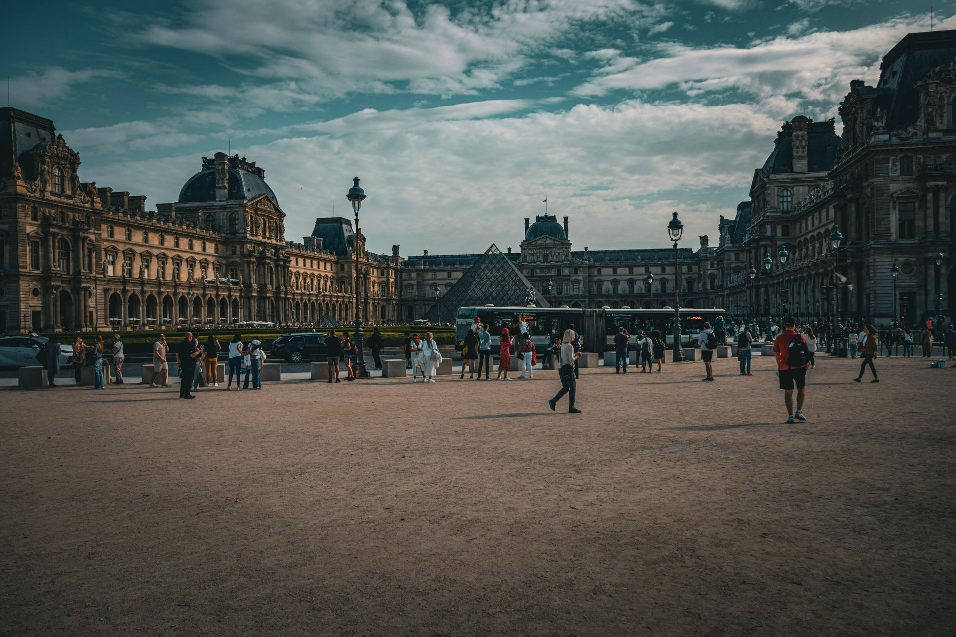 Large historic city square with people walking, flanked by ornate stone buildings under a cloudy sky