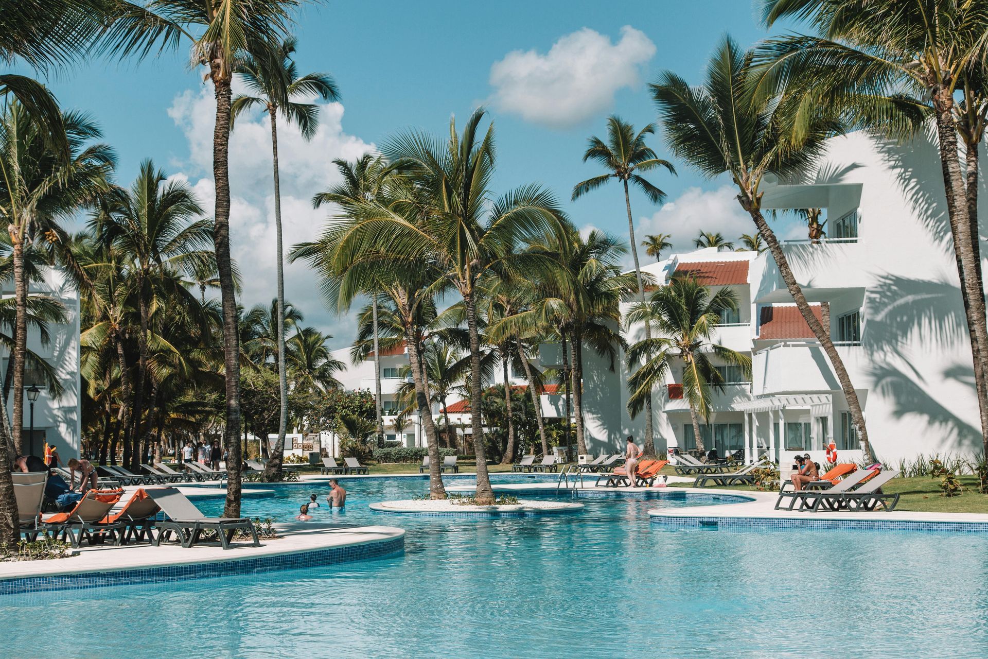 Tropical resort pool with palm trees, loungers, and a white building under a bright blue sky