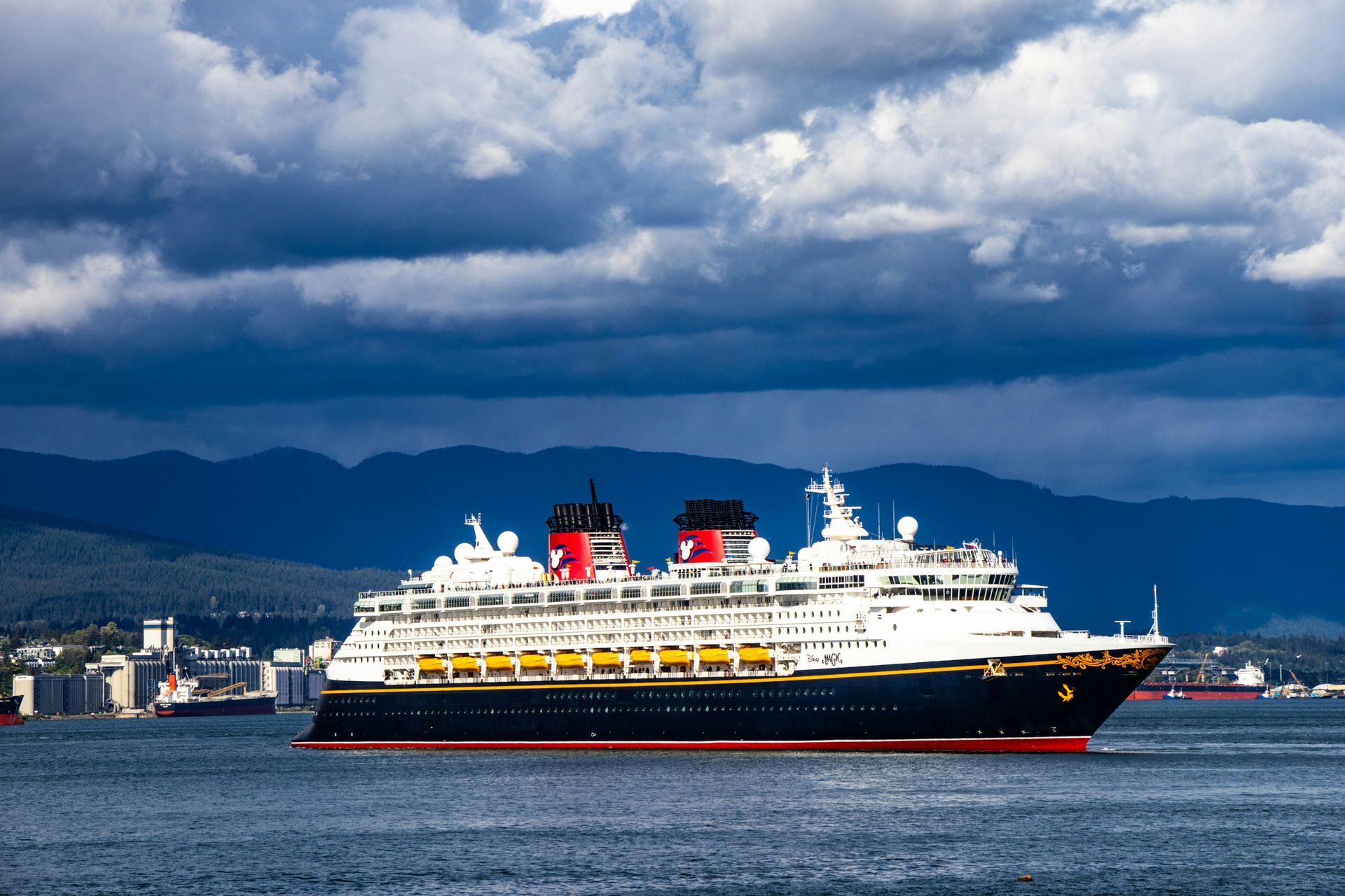 Large white cruise ship with red funnels sailing on calm water under cloudy mountains.