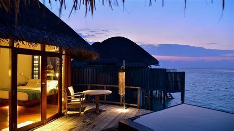 Sunset view of overwater resort bungalow with lit deck and lounge chairs beside the ocean