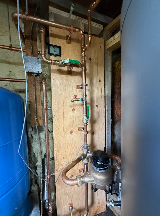 Utility room with water heater, blue tank, and exposed pipes against a plywood wall